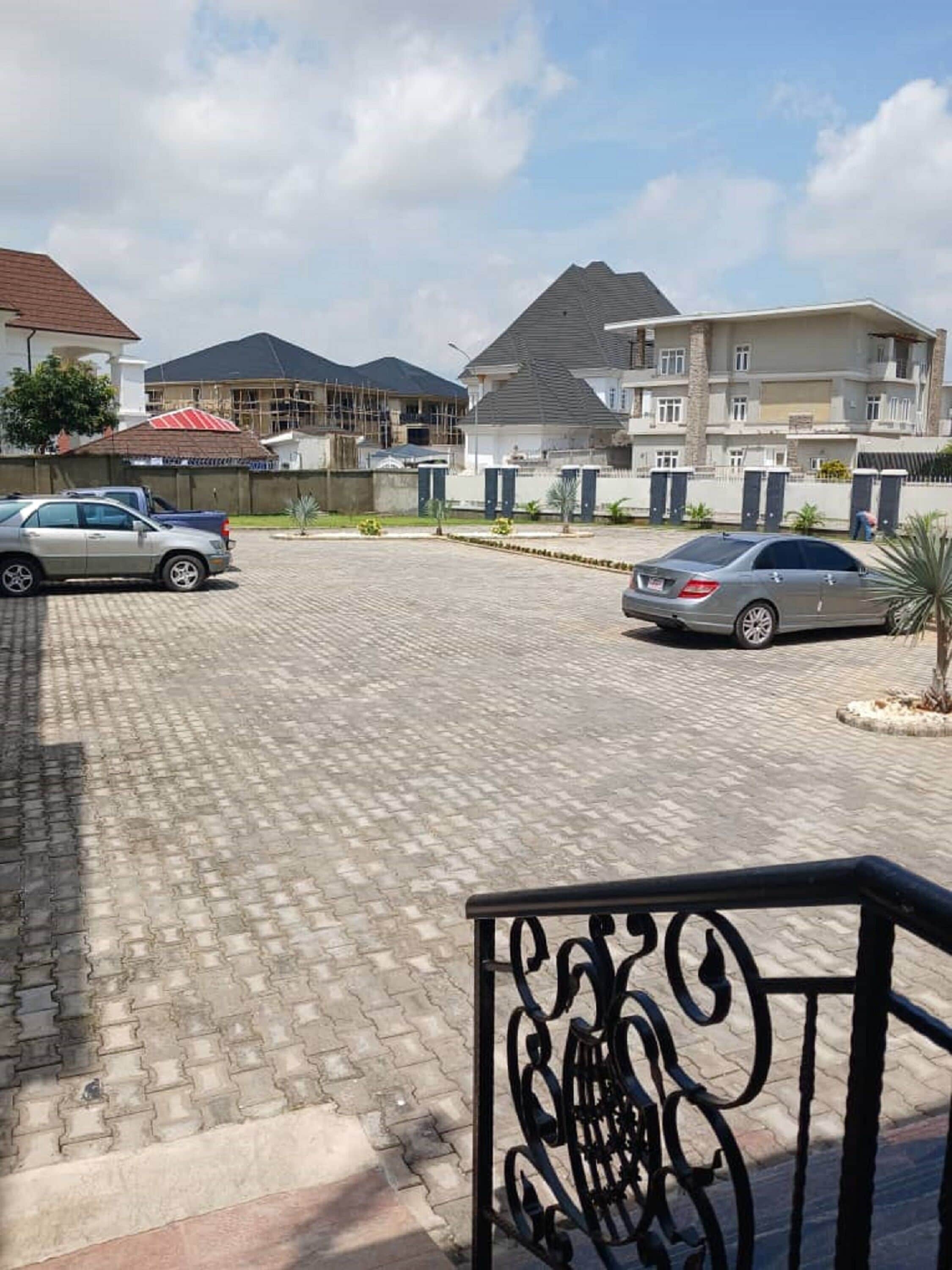 Une zone de stationnement dallée avec deux voitures, un bâtiment en construction et un immeuble d’habitation doté d’un toit en pente.