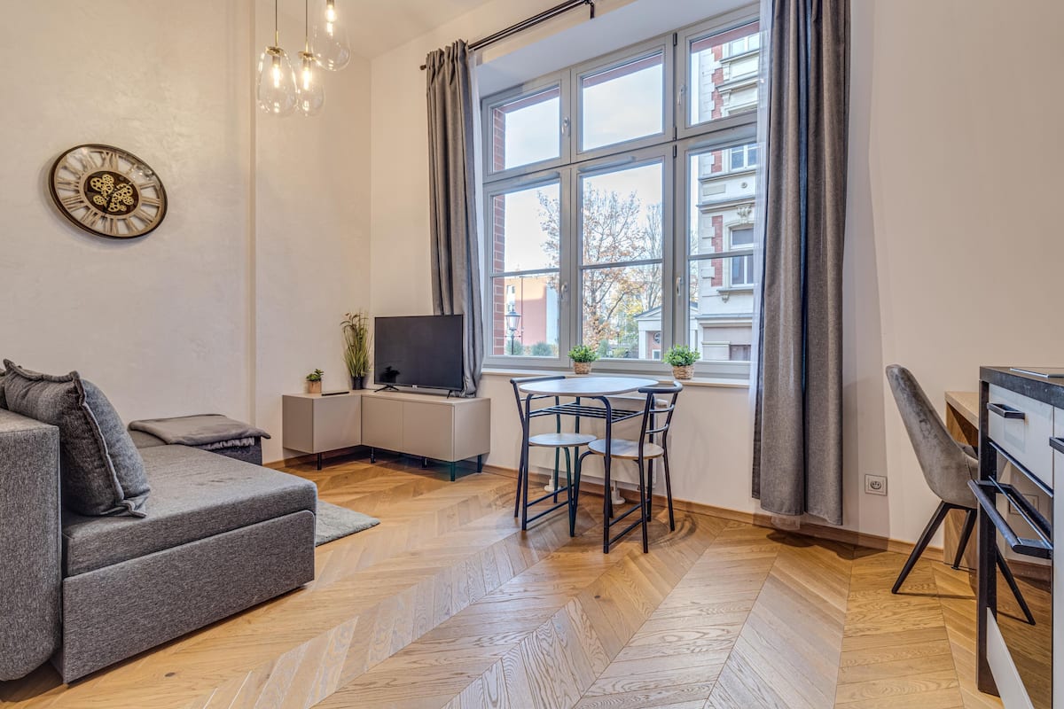 Ein modernes Wohnzimmer mit einer Couch, einem Fernseher, einem Esstisch mit Stühlen und einem Fenster mit Vorhängen.