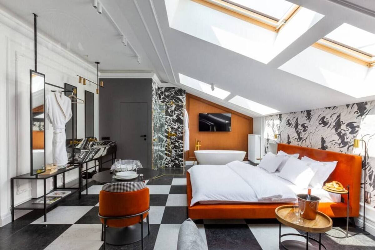 Une chambre d’hôtel moderne avec un sol à carreaux, un grand lit doté d’une tête de lit orange, une coiffeuse avec un miroir, une télévision et une lucarne.