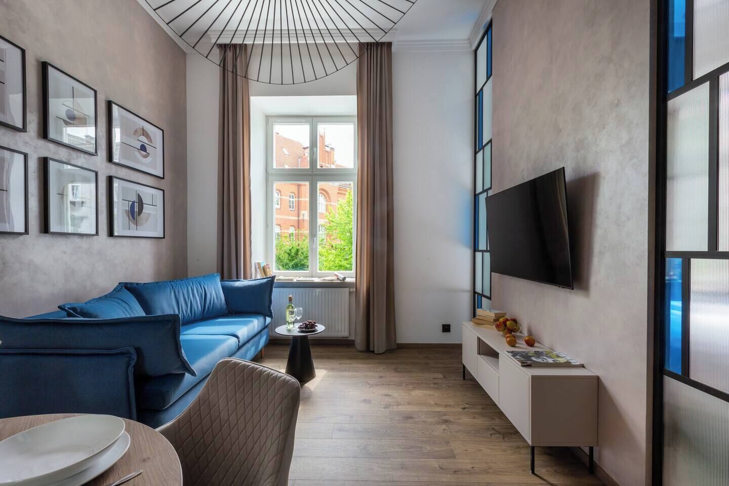 Un salon moderne avec un canapé bleu, une petite table ronde, un téléviseur fixé au mur et une fenêtre avec des rideaux.