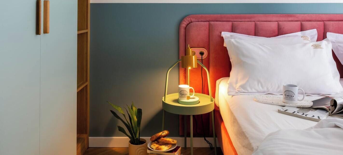 A bedroom with a bed, pillows, a bedside table with a lamp, a plant, and a small plate with bread.