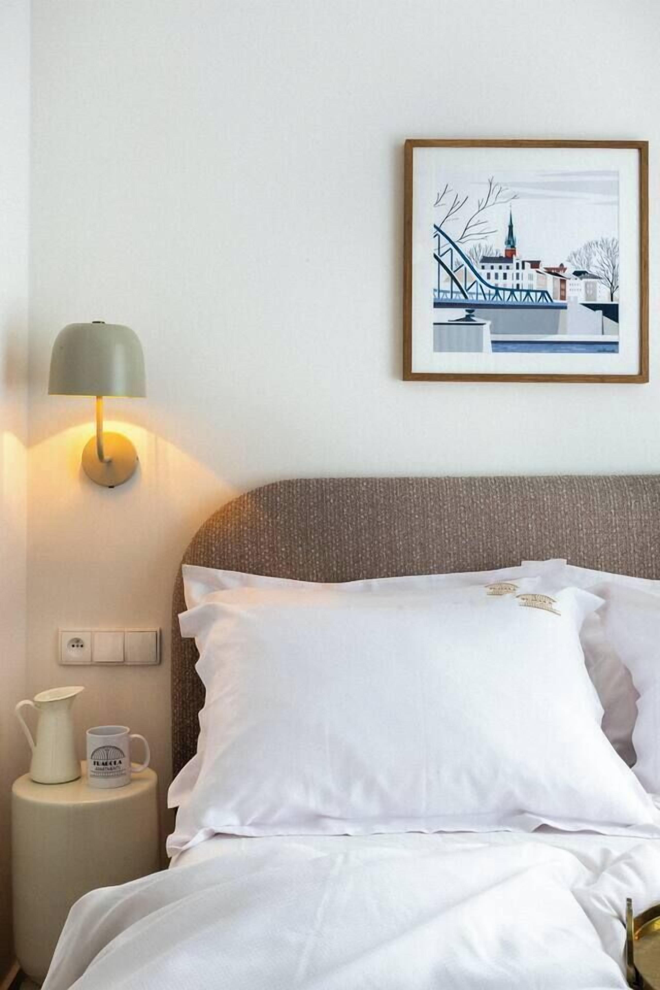 A neatly made bed with white linens, a wall-mounted lamp, a framed cityscape picture, and a small bedside table with a pitcher and mug.