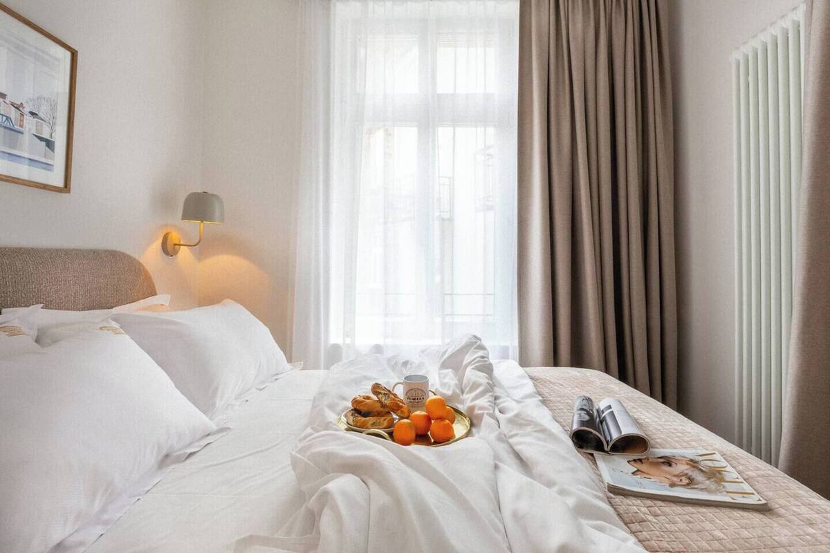 A hotel room with a bed, a bedside lamp, a window with curtains, and a tray with bread and oranges.