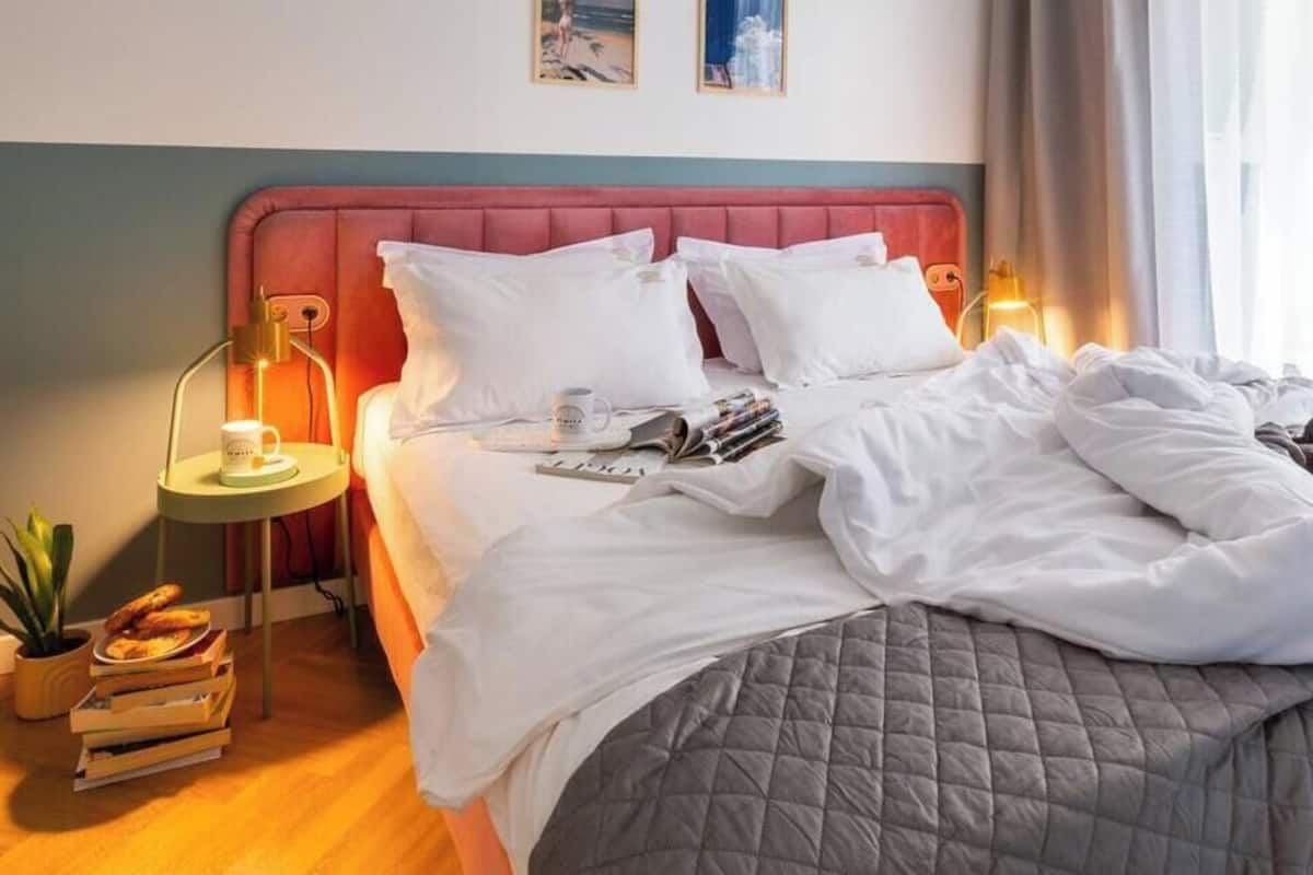 Un lit bien fait, avec des oreillers et une couverture, une table de chevet avec une lampe et une tasse, une petite plante et une fenêtre avec des rideaux.
