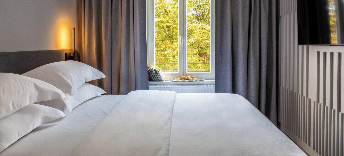 A bed with white linens, a window with curtains, a radiator, and a wall-mounted TV.