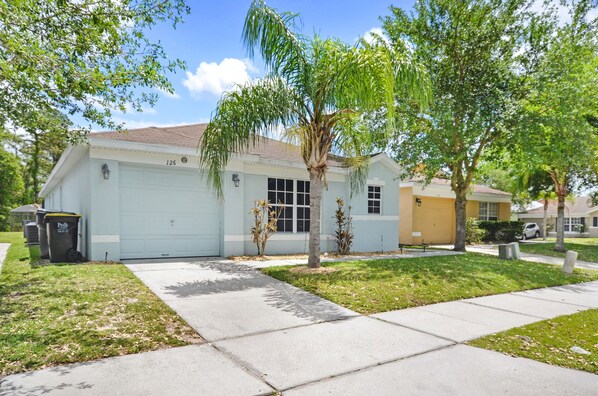 A single-story house with a garage, a small lawn, and a tree in front.
