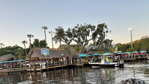 A docked boat with a thatched-roof building labeled "Gumbo's" in the background, surrounded by palm trees and a clear sky.