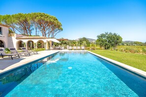 A rectangular swimming pool with clear blue water, surrounded by a white tiled deck. There are lounge chairs and a pergola with a white canopy.