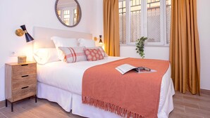 Una cama bien hecha con un edredón blanco y una manta roja, una lámpara de noche, un espejo y una ventana con cortinas.