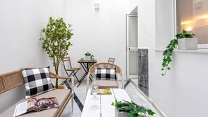 Un pequeño espacio exterior moderno con una mesa blanca, dos sillas de mimbre y un banco. Se ve una puerta de cristal que da a un baño, y una planta en maceta a la derecha.