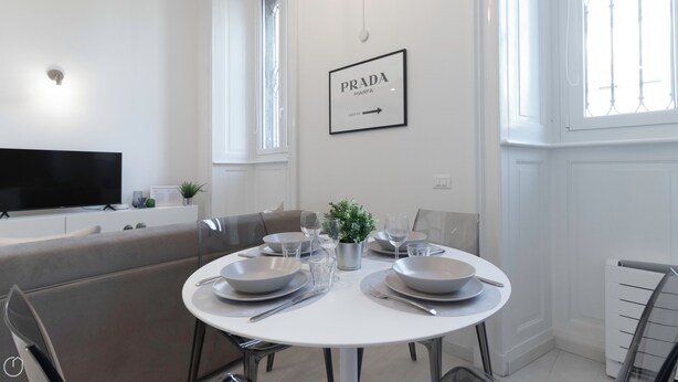 Un comedor moderno con una mesa blanca dispuesta para cuatro personas, un televisor sobre un mueble blanco, y un cartel enmarcado en la pared.