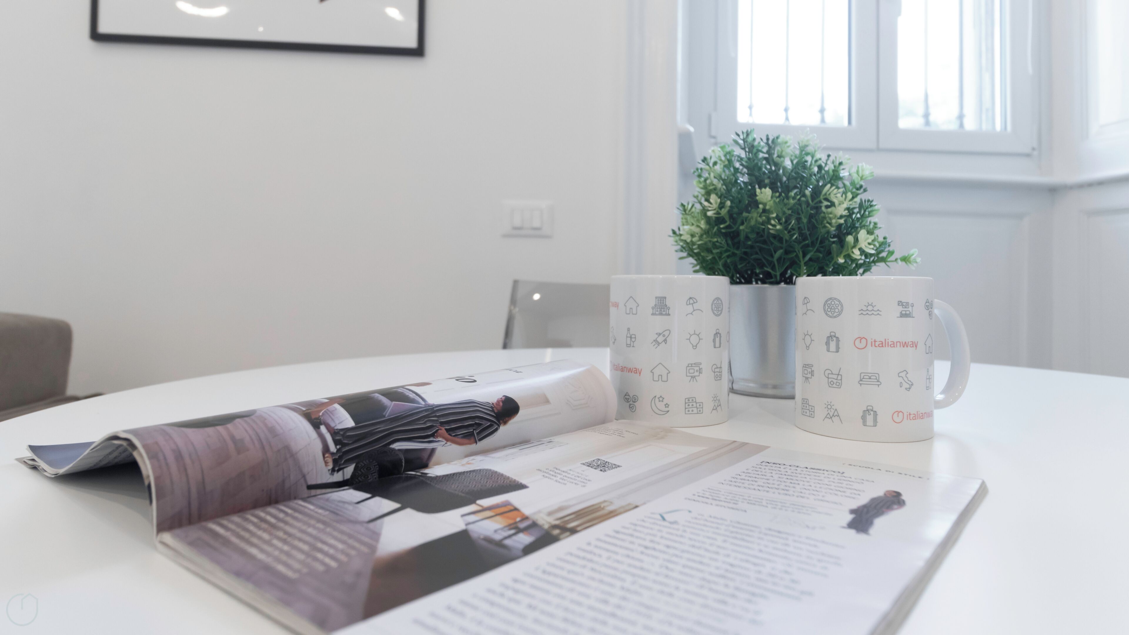 Een witte tafel met een tijdschrift, een mok met Italiaanse afbeeldingen en een kleine plant in een pot.