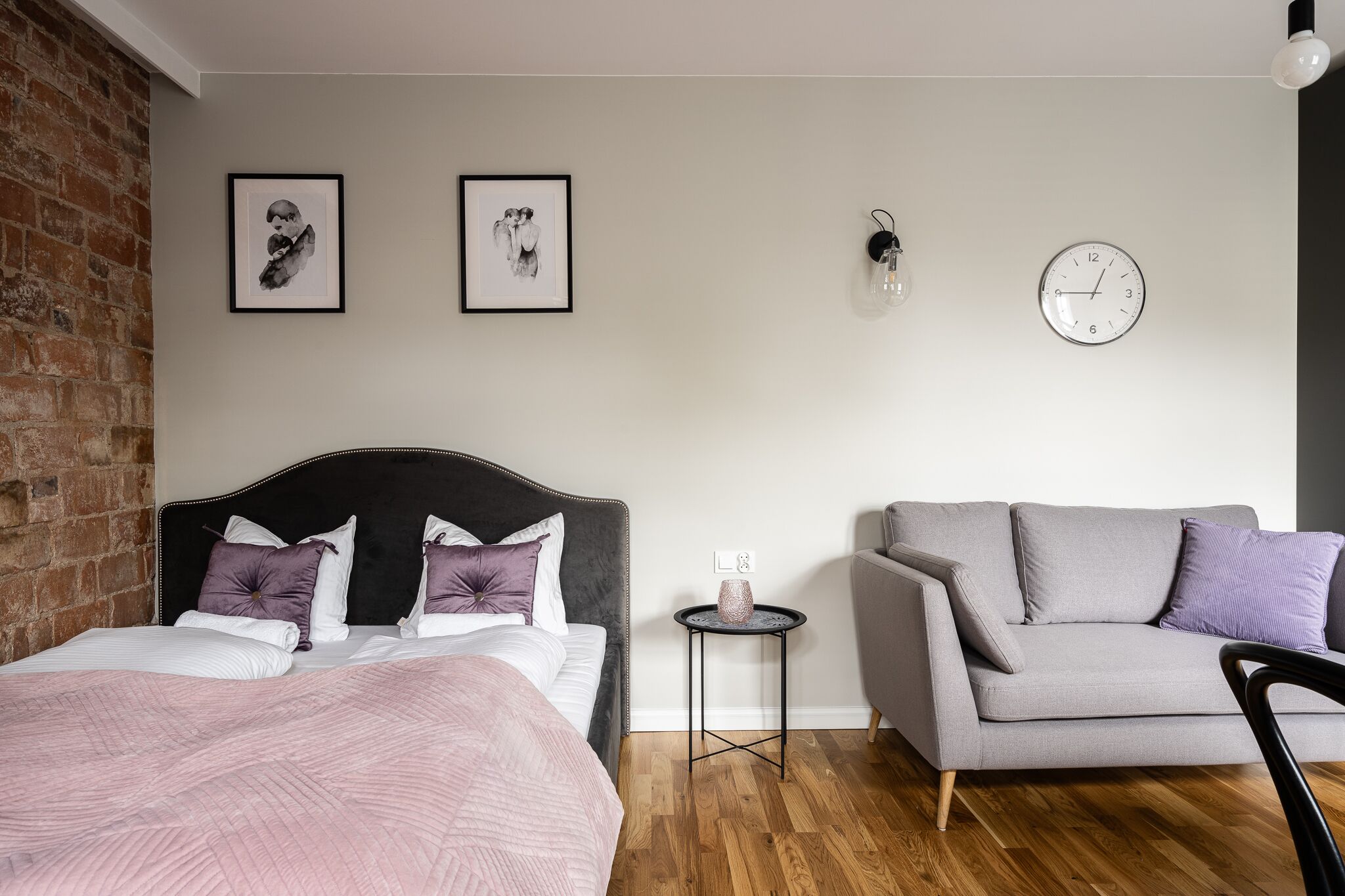 Un dormitorio con una cama, un sofá gris, una mesita auxiliar y dos cuadros enmarcados en la pared.
