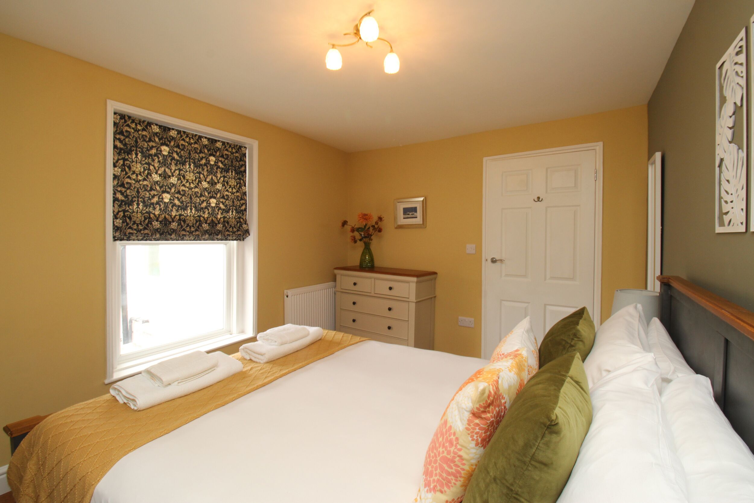 A bedroom with a bed, a window with a patterned blind, a dresser, and a door.
