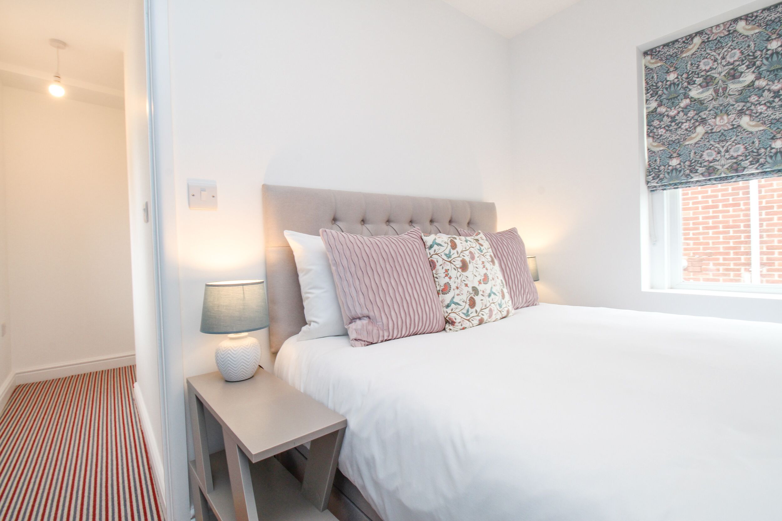 Une chambre à coucher avec un lit, une table de chevet avec une lampe, une fenêtre avec un store à motifs et un tapis rayé.