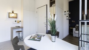 A modern dining area with a white table, chairs, and a vase with white flowers. There is a bookshelf with books and decorative items on the wall. - Italianway - Morosini 14 (Milan)
