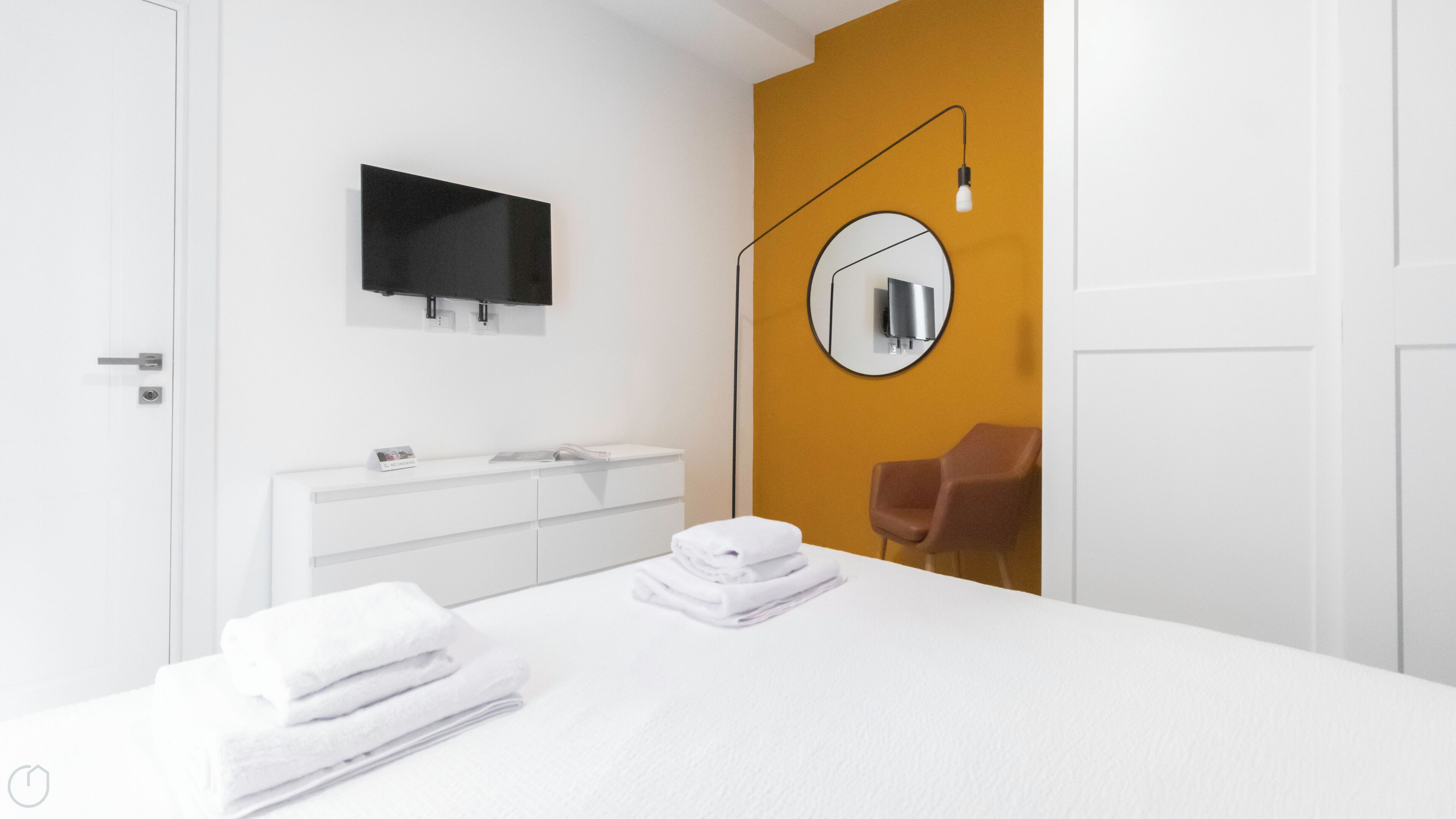 Una camera da letto moderna con un letto bianco, una cassettiera bianca, uno specchio rotondo, una televisione e una sedia marrone.