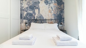 A neatly made bed with white linens and towels, a metal headboard, and two bedside lamps on a wall with a distressed paint finish.