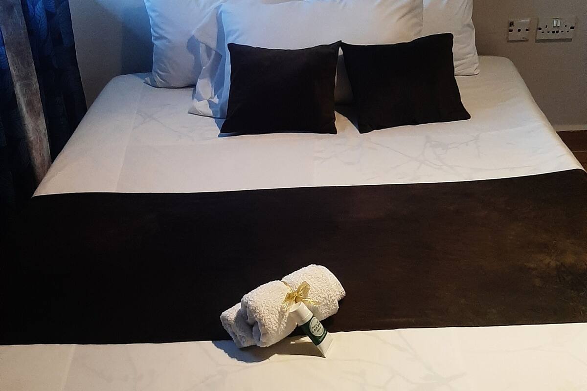 Ein ordentlich bezogenes Bett mit weißen und schwarzen Kissen, einem weißen Handtuch und einer Lotionflasche.