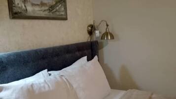 Un lit bien fait, avec du linge de lit blanc et une tête de lit sombre. Un tableau représentant un paysage, encadré, est accroché au mur au-dessus du lit.