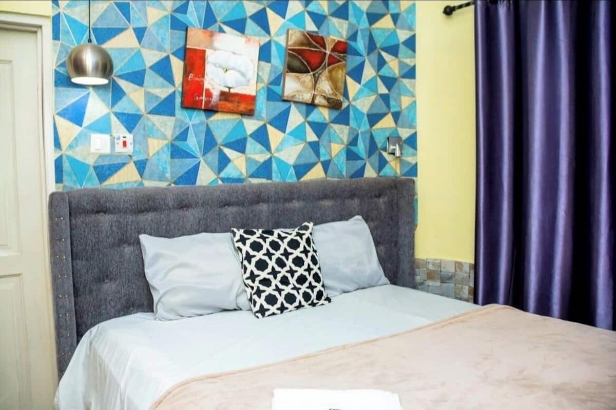 Ein Bett mit grauem Kopfteil, einem gemusterten Kopfkissen und einer blauen geometrischen Wand.