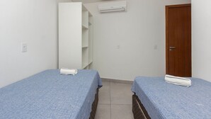 Une petite chambre propre, avec deux lits simples, une porte en bois, une armoire blanche et un climatiseur fixé au mur.