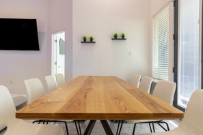Un salón de conferencias con una gran mesa de madera, sillas blancas, dos estanterías con plantas y un televisor en la pared.