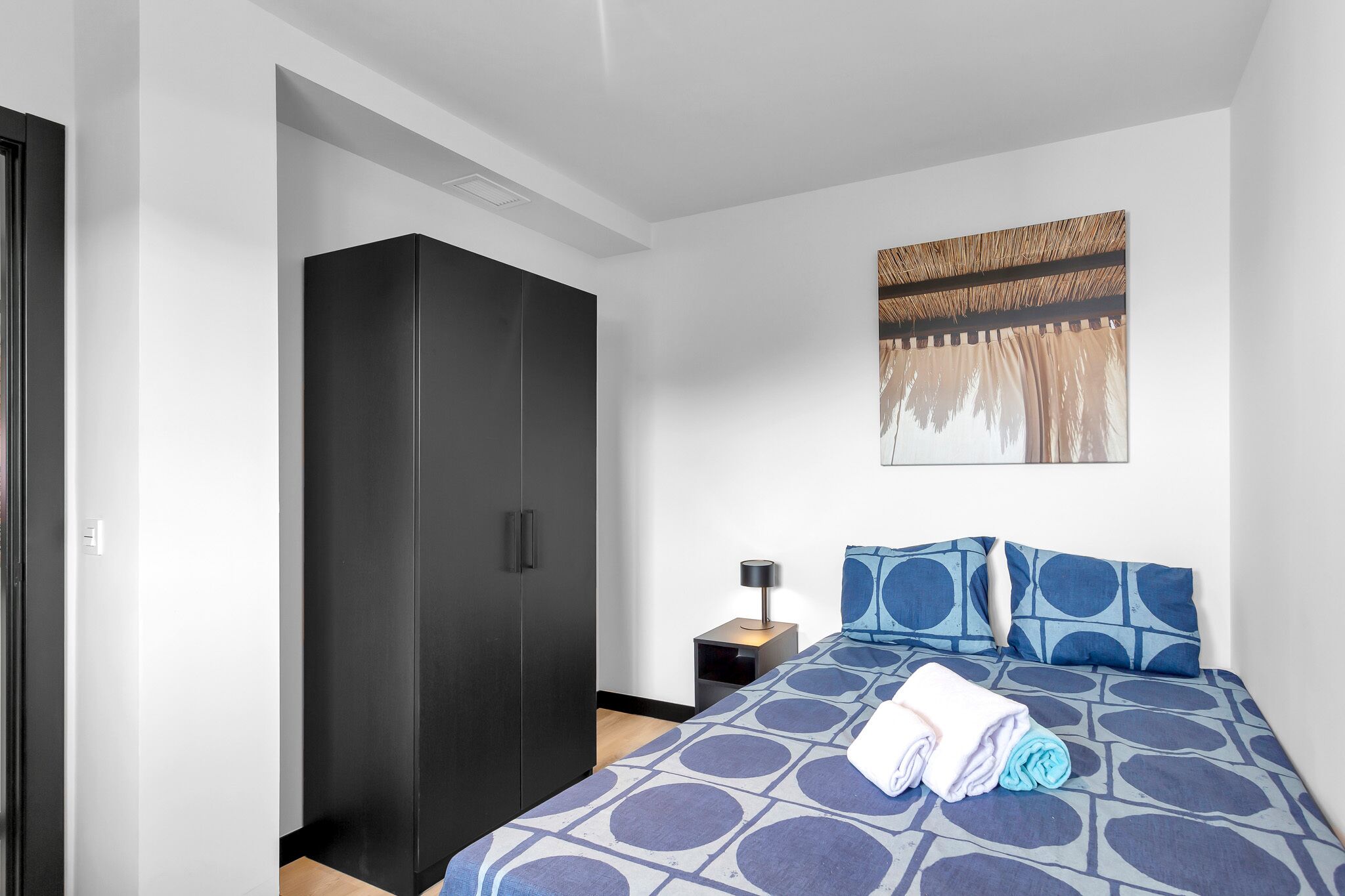 A hotel room with a bed, a wardrobe, a nightstand, and a wall-mounted artwork.
