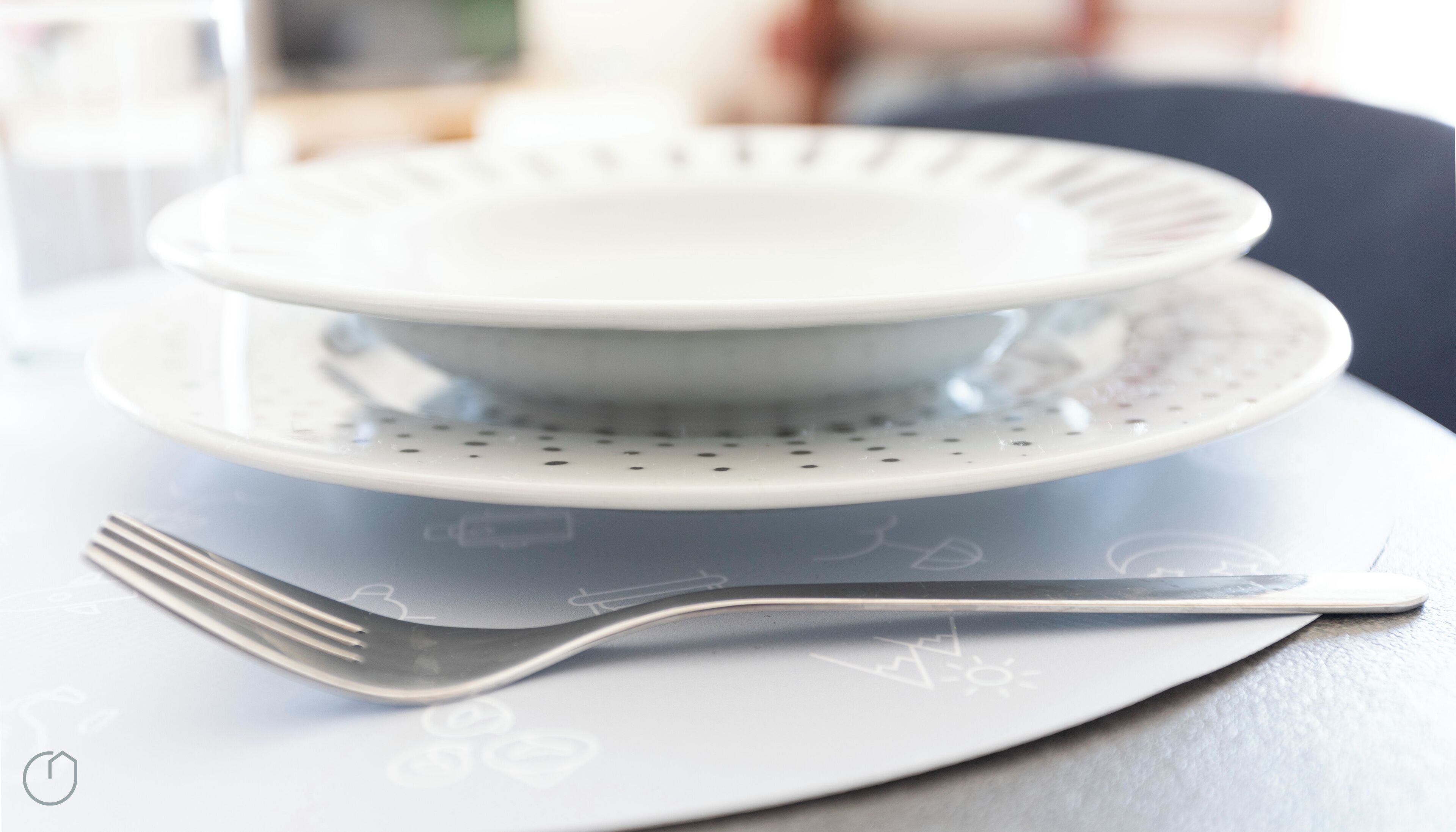Un plato blanco con un bol encima, colocado sobre una mesa blanca, con un tenedor a un lado.
