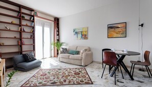 A living room with a sofa, a coffee table, and a dining table with chairs. There is a bookshelf filled with books and decorative items. A painting hangs on the wall, and a potted plant is on the floor. - Italianway Easy - Sirte 6 (Milan)