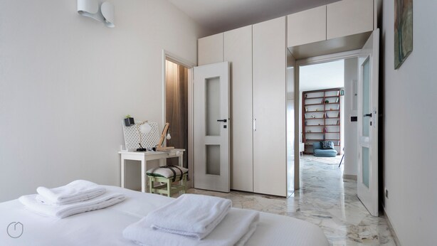 Een moderne slaapkamer met een bed, witte handdoeken, een bureau met lamp, een boekenkast en een deur die naar een andere kamer leidt.