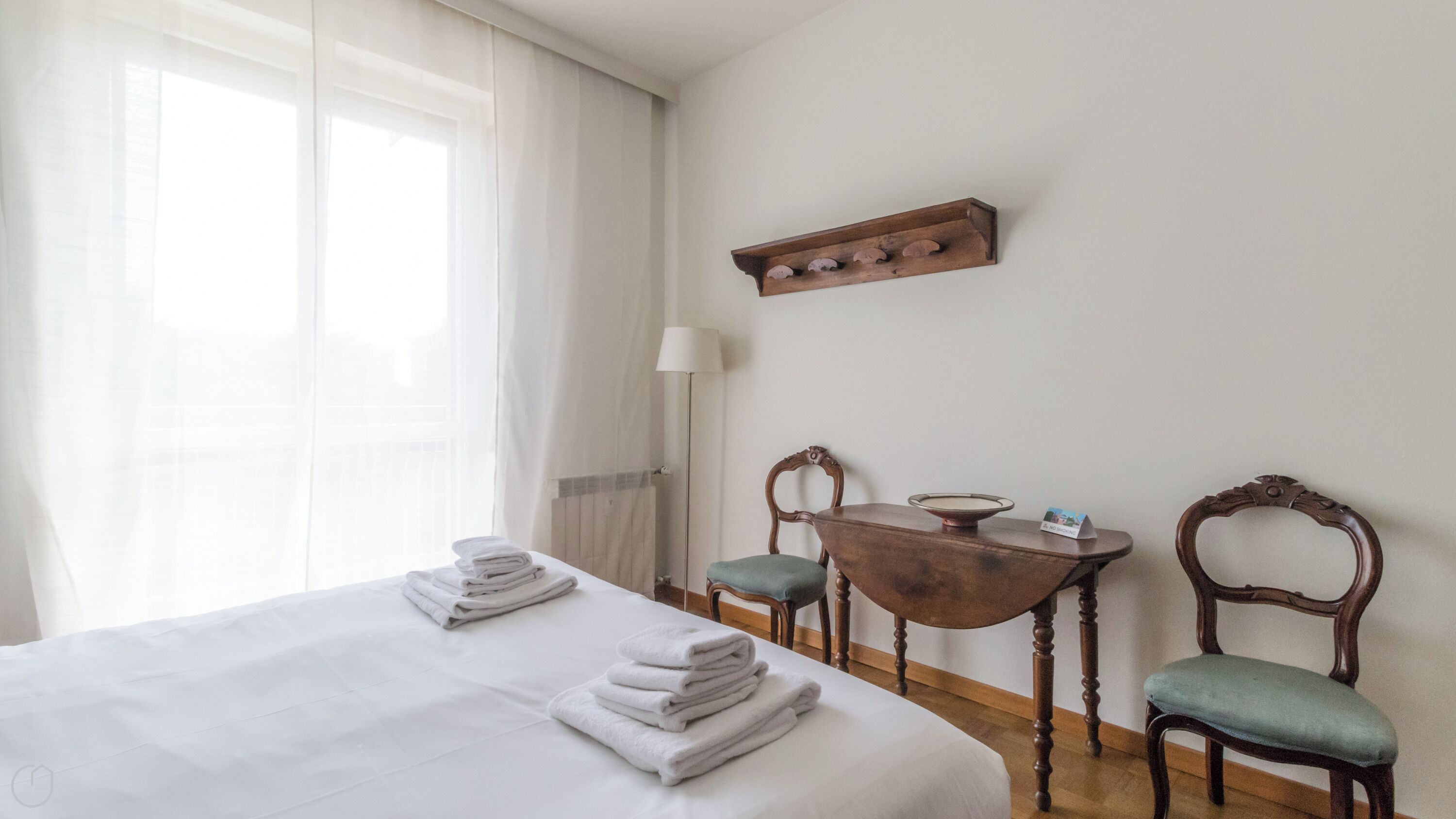 Siisti hotellihuone, jossa on huolellisesti pedattu sänky, pieni puinen pöytä, jossa on kulho ja nimikortti, kaksi pehmustettua tuolia ja lattiavalaisin.
