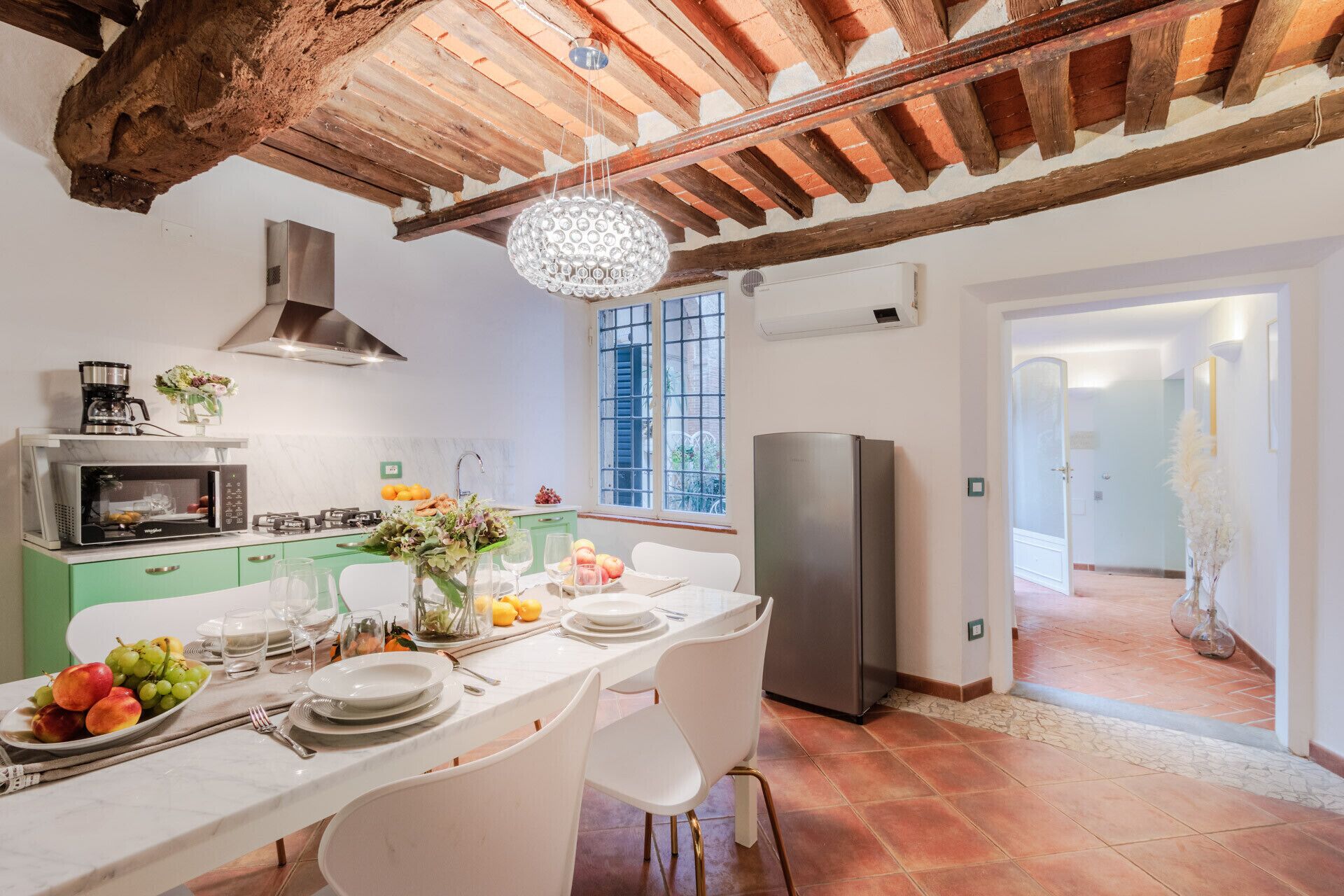 A kitchen with a dining area, featuring a wooden ceiling, a stainless steel refrigerator, and a dining table set with plates, glasses, and fruit.