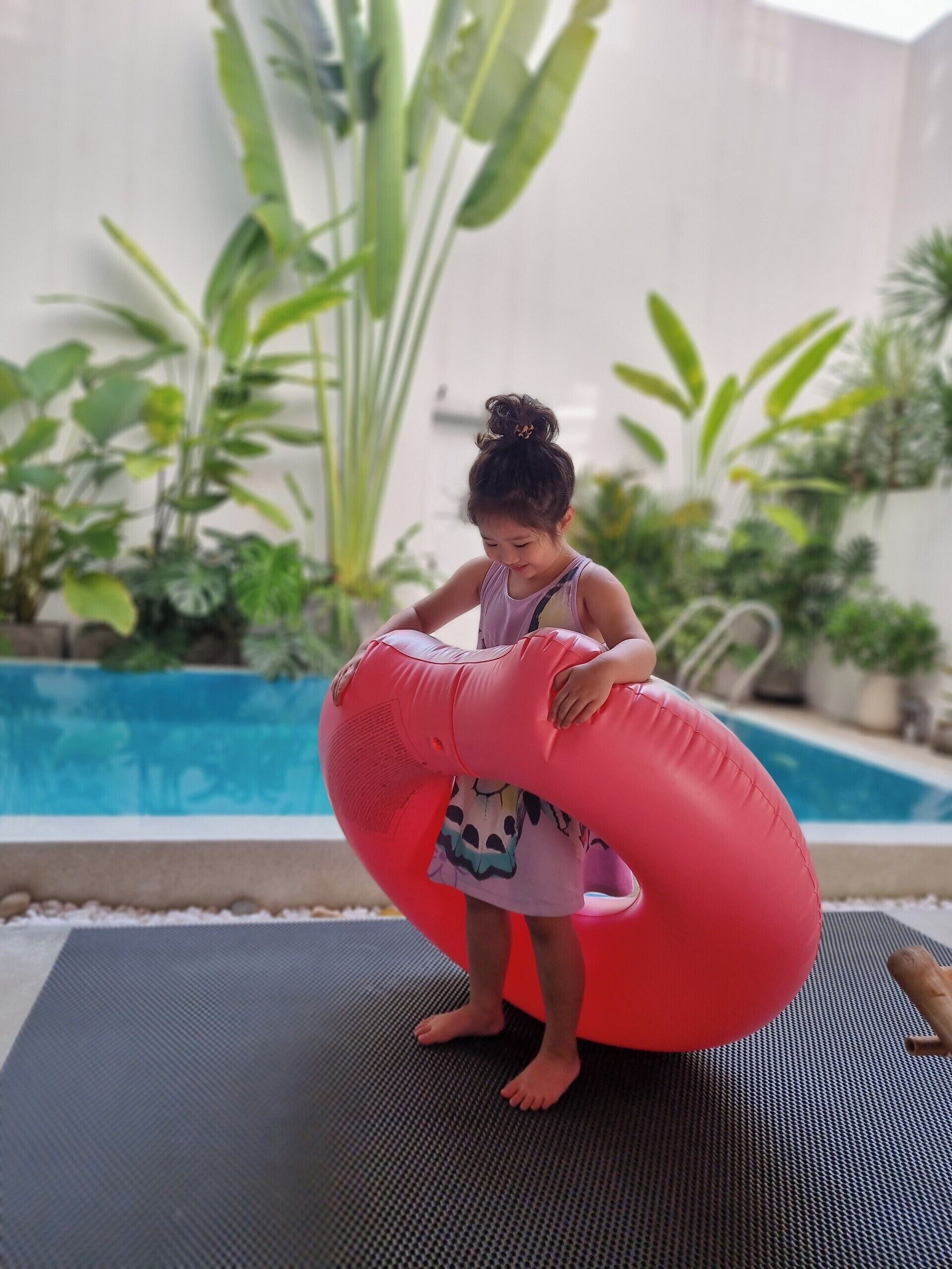 A child playing with a red inflatable ring near a pool with a black mat on the ground.