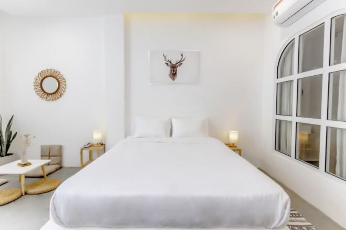 Habitación de hotel moderna con una cama grande, un espejo redondo, una cabeza de ciervo enmarcada, una mesita de noche con una lámpara y un ventanal amplio.
