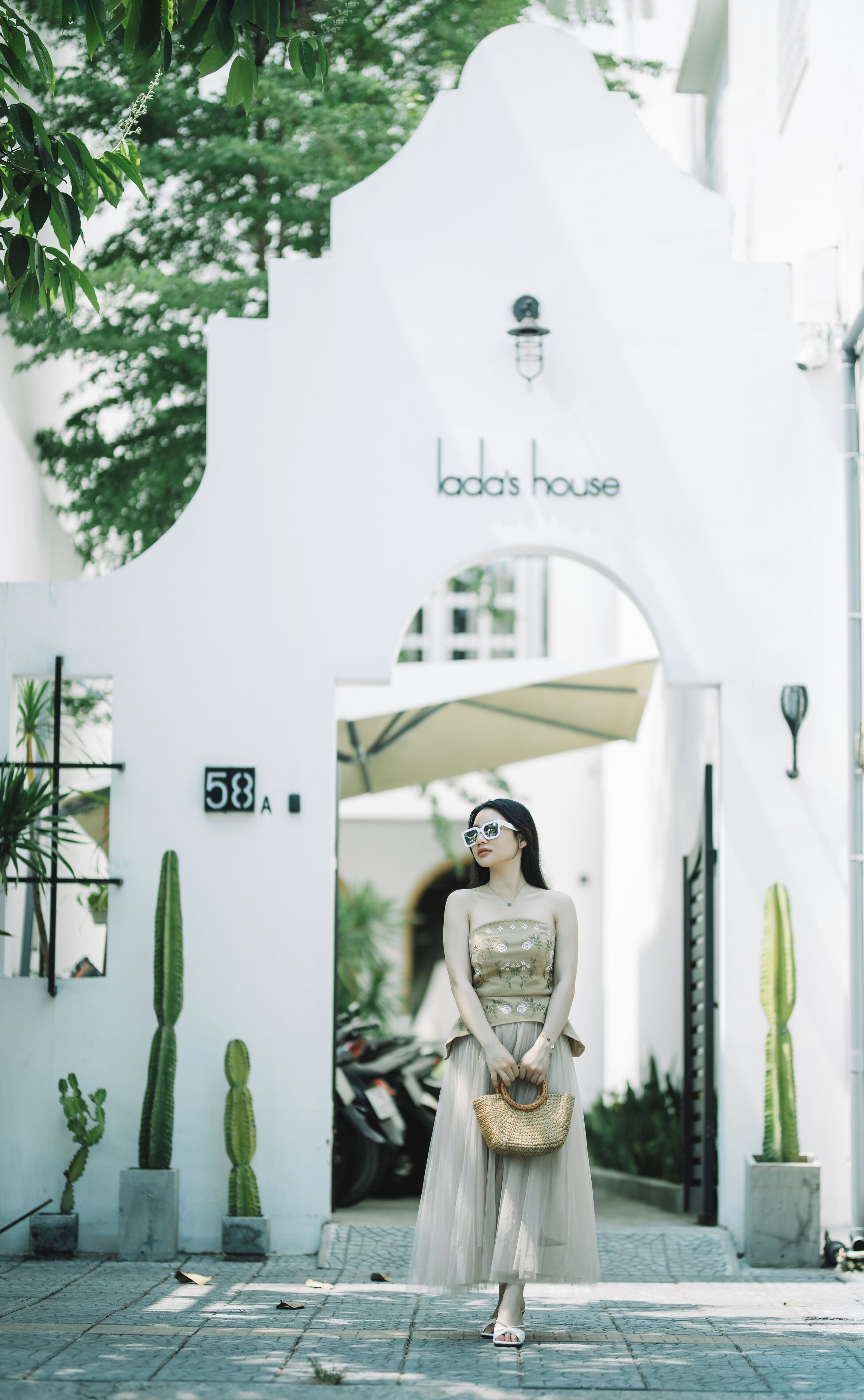 A woman standing in front of a white building with a sign reading "Lada's House" and the number "58" on the wall.