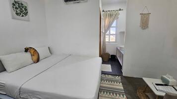 A hotel room with a bed, a desk, a chair, a window with curtains, an air conditioning unit, and a wall-mounted artwork.
