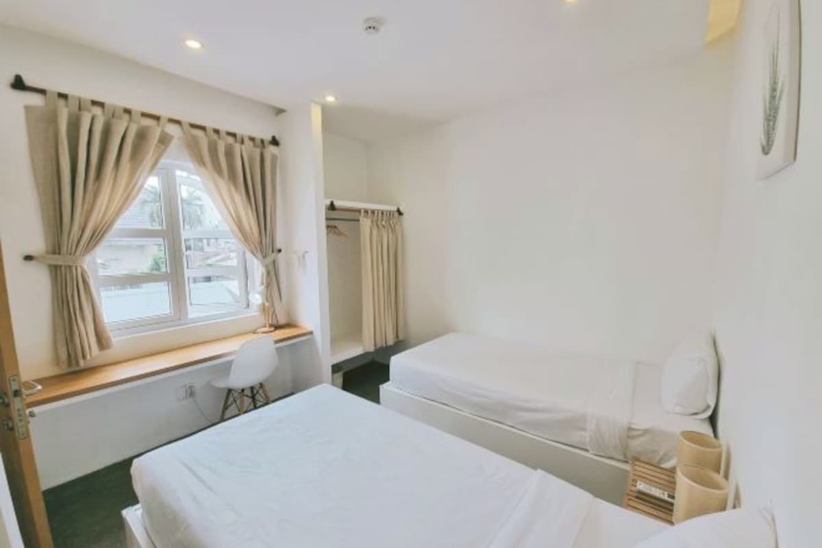 Habitación de hotel con dos camas, una ventana con cortinas, una silla y una mesita con lámpara.
