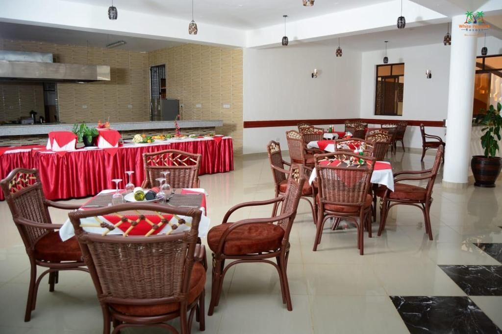 A dining area with wicker chairs and tables, a kitchen in the background, and a white wall with a red stripe.