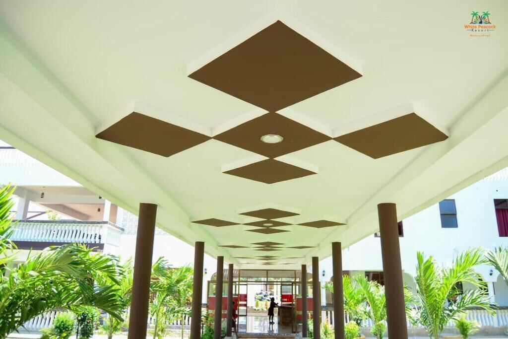 Un paseo techado con un diseño geométrico en el techo, palmeras y un edificio al fondo.