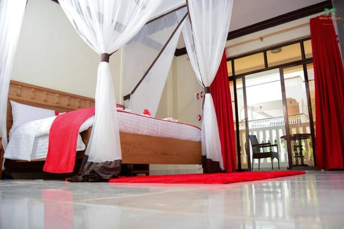 Quarto com cama, dossel branco, cortinas vermelhas e vista para o exterior.