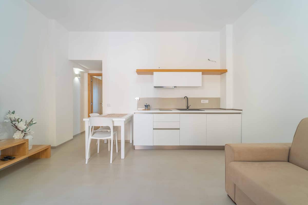 Una cocina moderna con armarios blancos, encimera de madera y un lavamanos empotrado. Hay una mesa de comedor blanca con dos sillas y un sofá beige en la zona de estar.