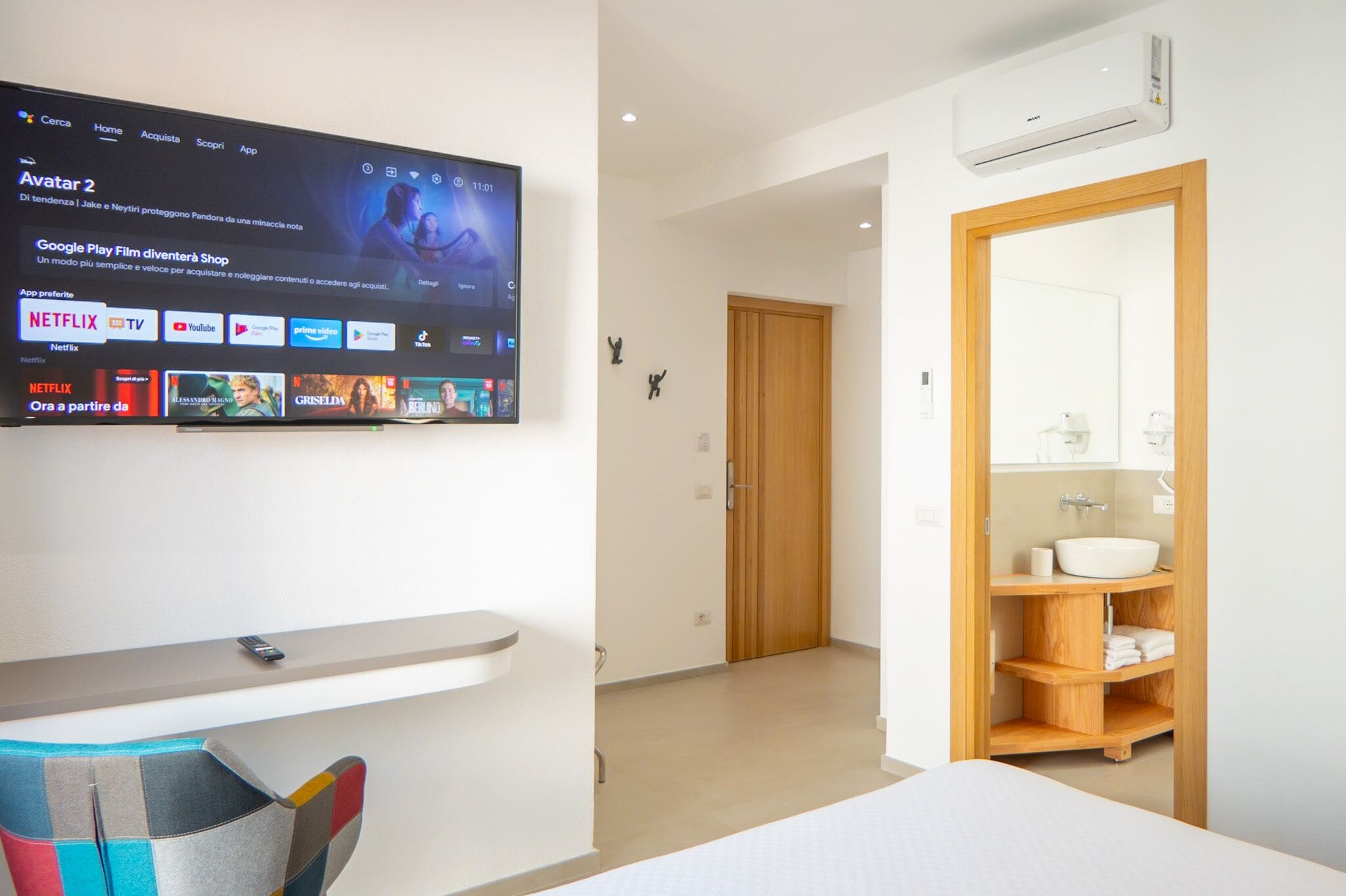 Ein modernes Hotelzimmer mit einem an der Wand befestigten Fernseher, der Streamingdienste anzeigt, einer Holztür, einem Badezimmer mit Waschbecken und Spiegel sowie einem kleinen Regal.