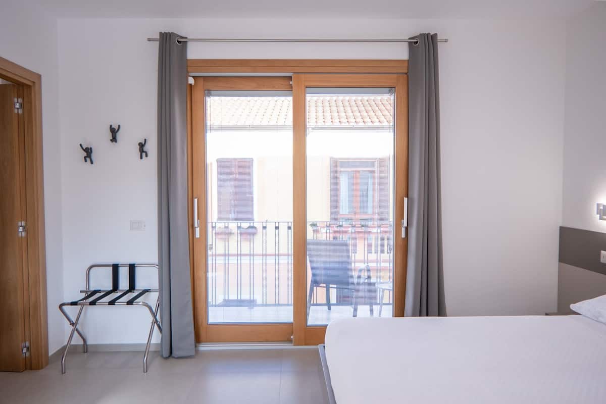 Una habitación de hotel con una cama, un tendedero, un balcón con vistas a edificios y una puerta.