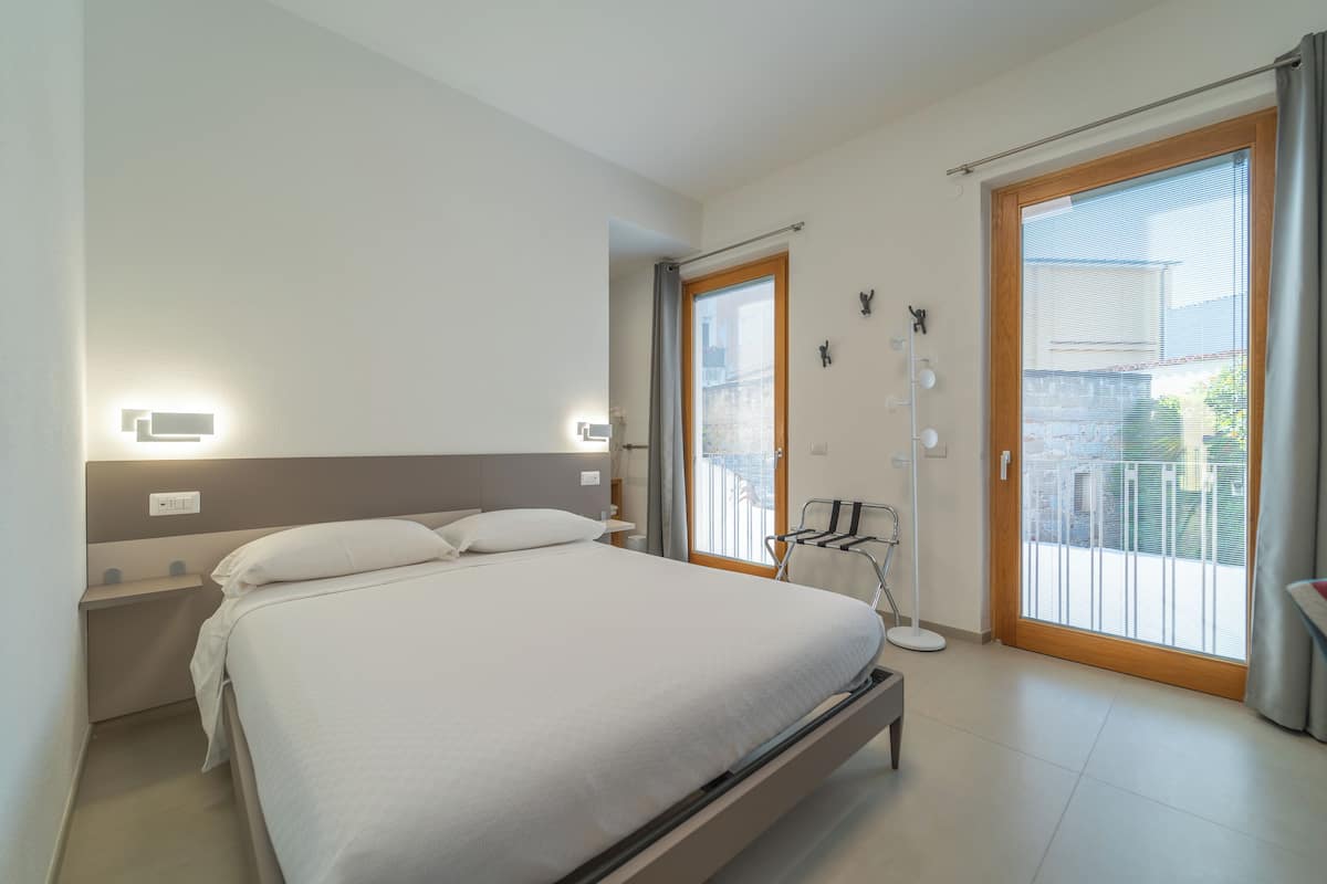 Una habitación de hotel moderna con una cama grande, una mesita de noche, una silla, una ventana con vistas y una puerta corredera al balcón.