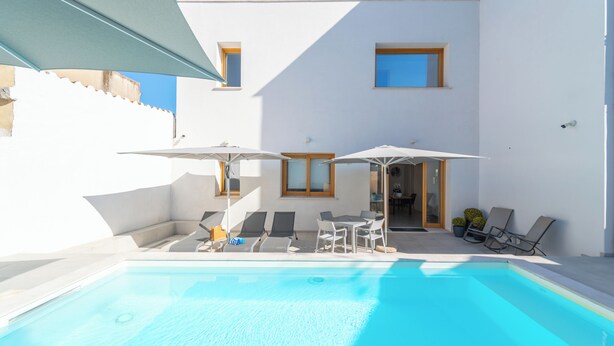 Un área de piscina exterior moderna con sillones de descanso y sombrillas, un cielo azul despejado y un edificio blanco con ventanas.