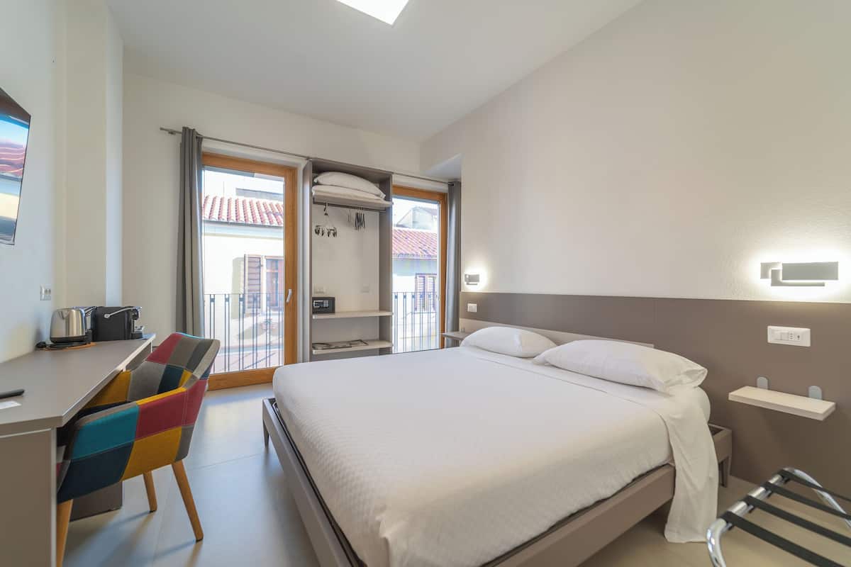 Habitación de hotel moderna con cama, escritorio, una silla colorida, balcón con vistas y una kitchenette pequeña.