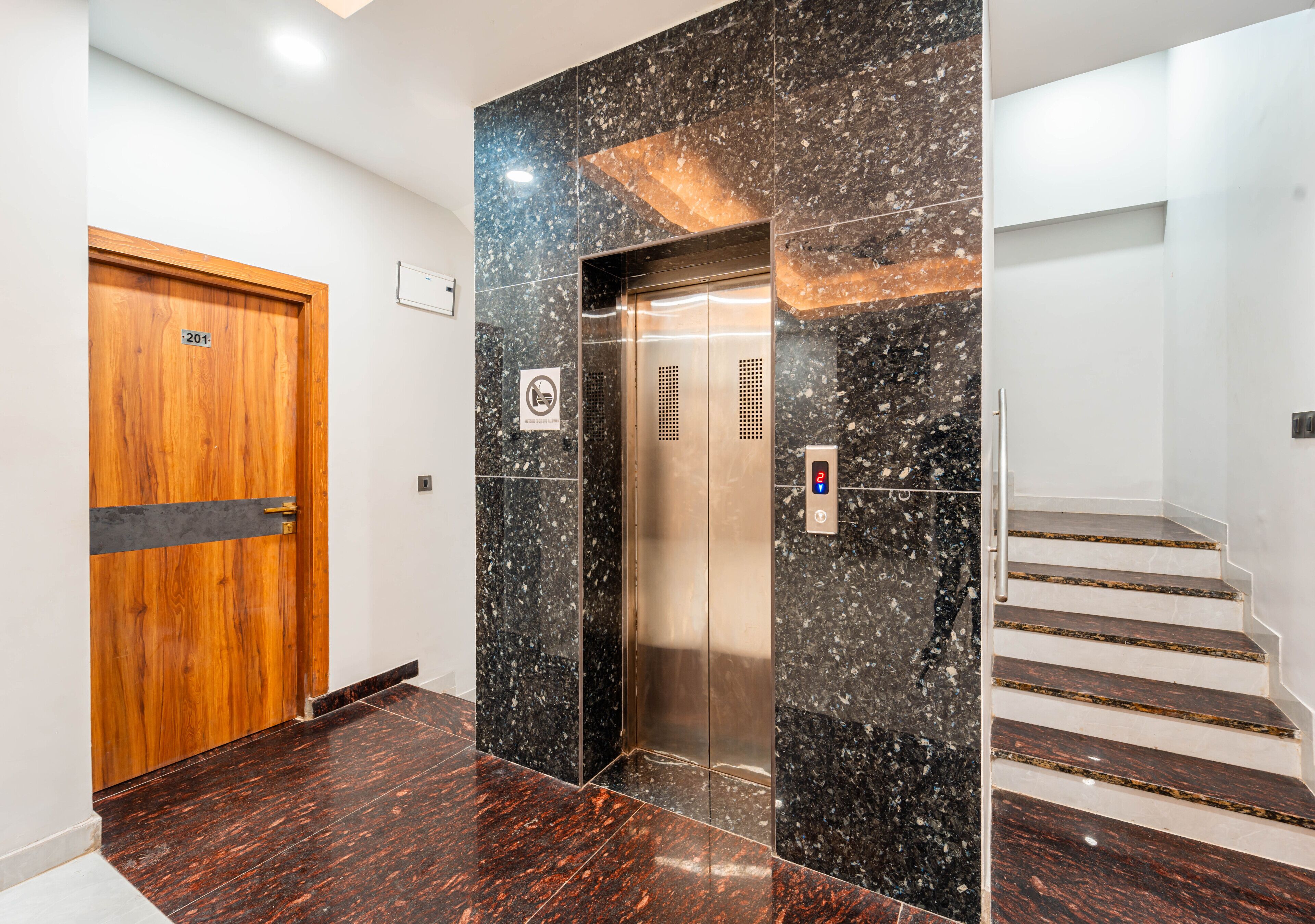 An elevator with a digital control panel, a wooden door with a number plate, and a staircase with marble steps.