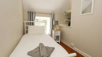 Una habitación pequeña con cama individual, suelo de madera, mesita de noche con lámpara, espejo y ventana con cortinas.