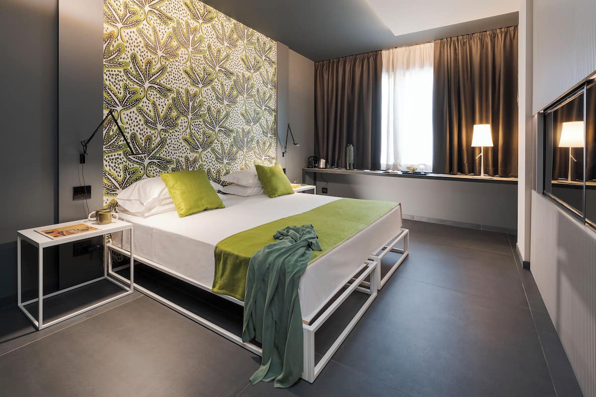 A modern hotel room with a large bed, green pillows, a bedside table with a lamp, and a window with curtains.